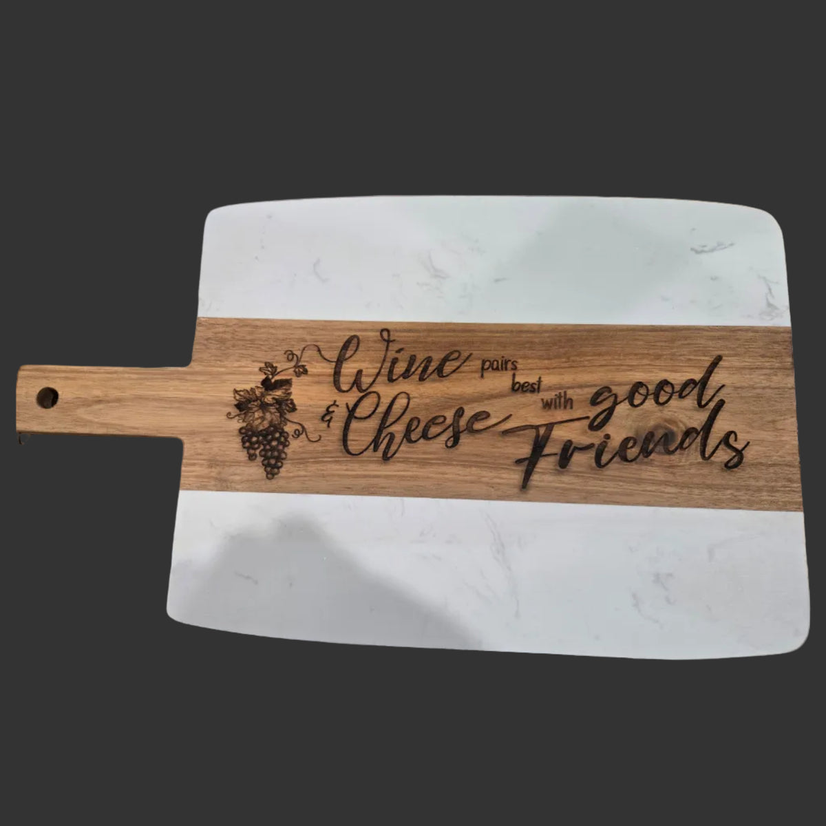 A rectangular wooden serving board with a natural wood handle and white stone or composite end panels, engraved with the phrase 'Wine pairs best with good friends' and a grapevine illustration. The board is photographed on a dark wood tabletop.